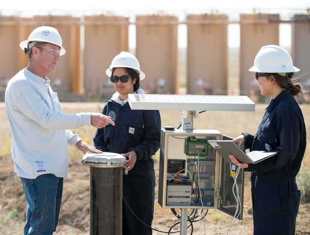 Three people wearing hard hats around a water level monitoring well site
